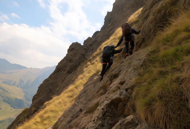 Two climbers ascend a steep, rocky path in a mountainous landscape